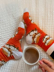 Someone wearing a pair of fall-themed socks with orange and white color and fox prints and holding a cup of coffee.