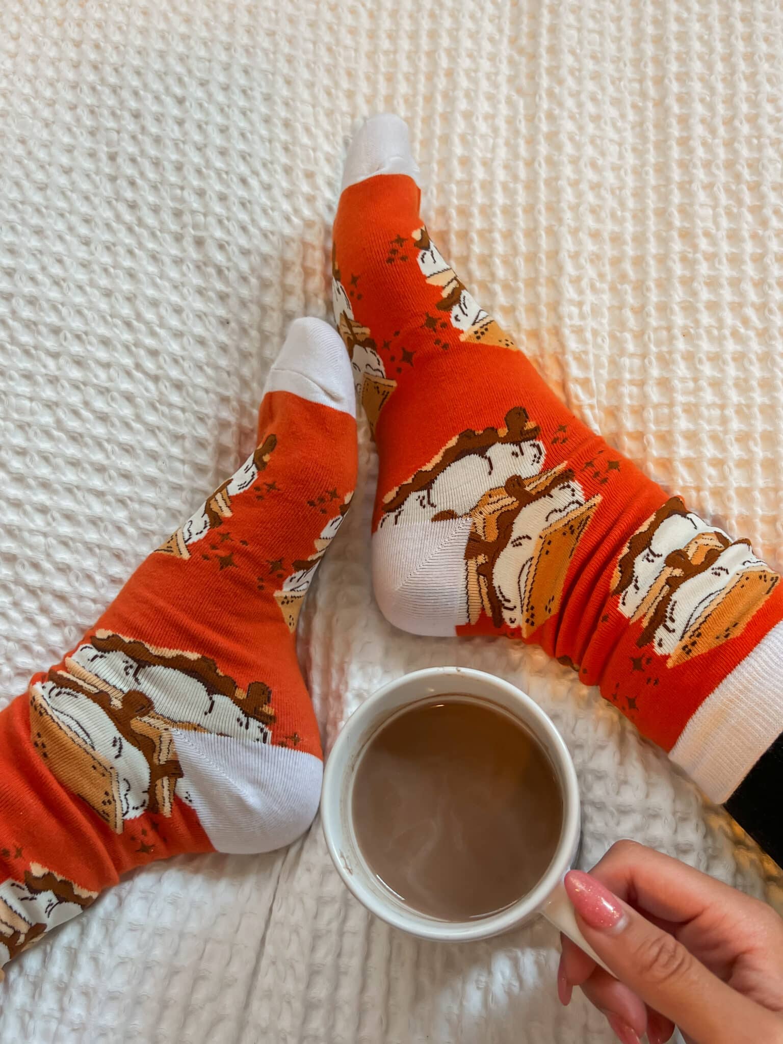 Someone wearing a pair of fall-themed socks with orange and white color and fox prints and holding a cup of coffee.