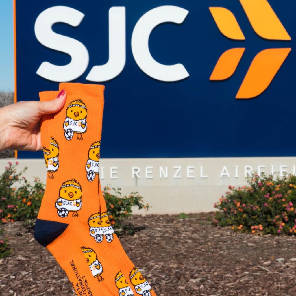customer holding up a pair of custom branded socks for SJC company in front of the establishment, the socks feature a branded orange backcground with cute sporting chicken graphics