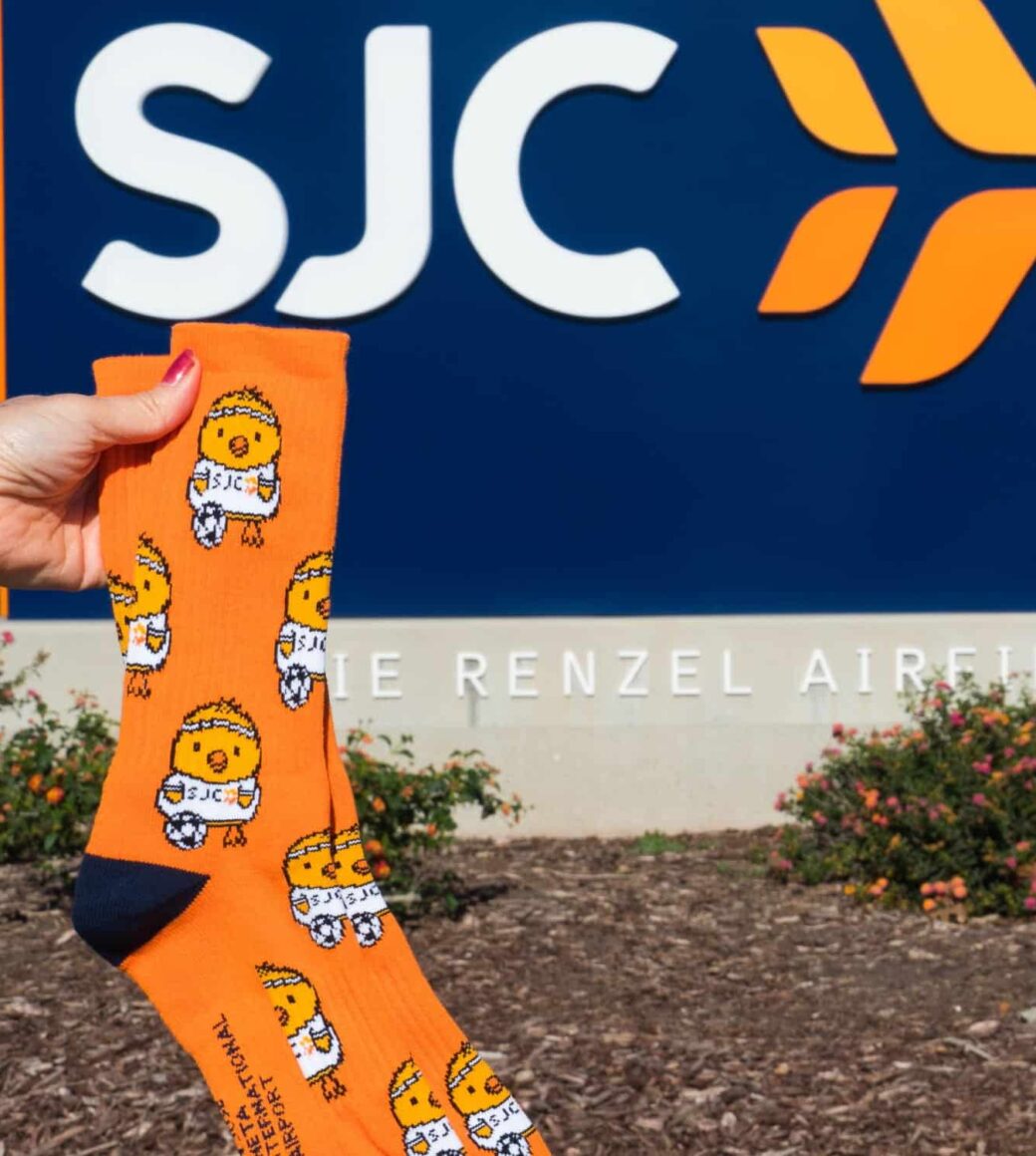 customer holding up a pair of custom branded socks for SJC company in front of the establishment, the socks feature a branded orange backcground with cute sporting chicken graphics